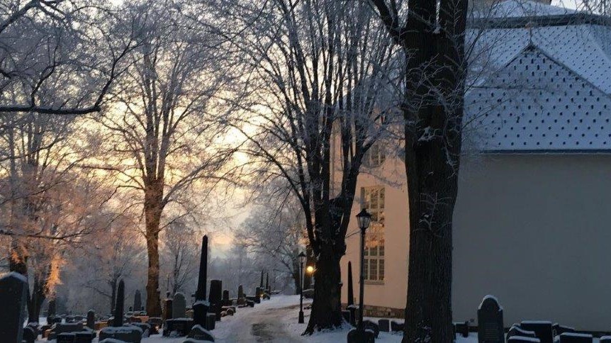 Et vinterbilde av en kirkegård i Hamar, der gravsteiner er omgitt av snødekte trær og en rolig atmosfære. Kveldssolen skaper et varmt skjær i bakgrunnen, et bygg står til venstre med frostede detaljer på taket. Foto.